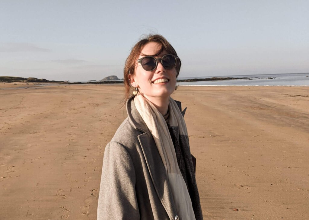 Me (Oonagh) smiling on a beach in a long grey felt coat and my hands in my pockets